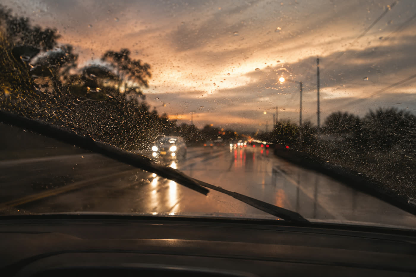 Blick durch regennasse Windschutzscheibe mit Scheibenwischer auf blendende Scheinwerfer in der Dämmerung